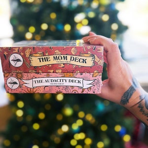May include: Two pink and white card decks with illustrated designs. The top deck says "The Mom Deck - The Pine Torc Shop" and the bottom deck says "The Audacity Deck - The Pine Torc Shop".