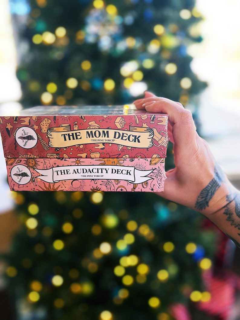 May include: Two pink and white card decks with illustrated designs. The top deck says "The Mom Deck - The Pine Torc Shop" and the bottom deck says "The Audacity Deck - The Pine Torc Shop".