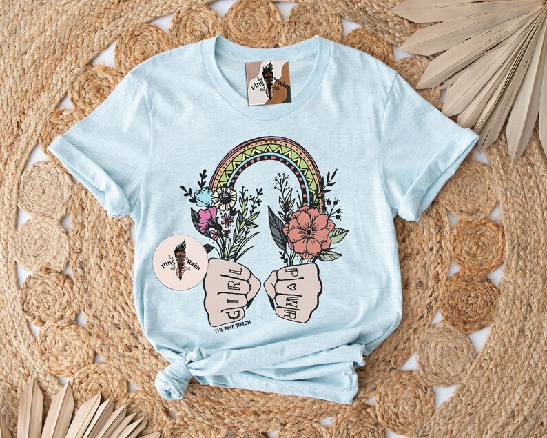 May include: Light blue t-shirt with a rainbow, flowers, and two fists with the words "Girl Power" and "The Pink Torch".
