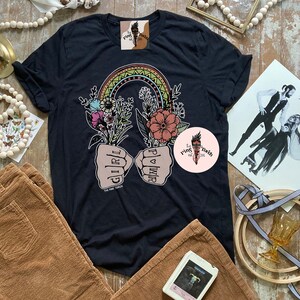 May include: Black t-shirt with a rainbow arch and two fists with the words "Girl Power" and "Girl" written on them. The fists are holding flowers. The text "The Pink Torch Est. 2018" is at the bottom of the shirt.