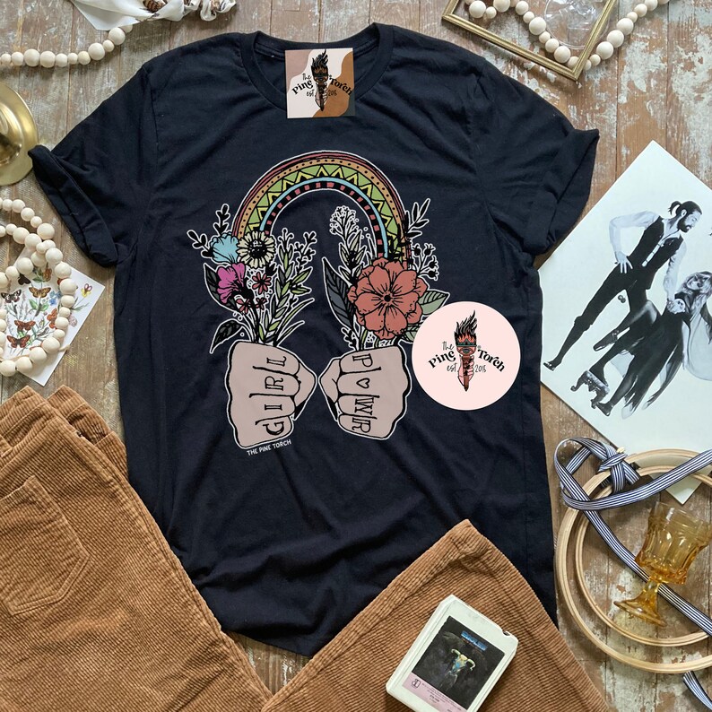 May include: Black t-shirt with a rainbow arch and two fists with the words "Girl Power" and "Girl" written on them. The fists are holding flowers. The text "The Pink Torch Est. 2018" is at the bottom of the shirt.