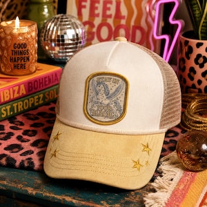 May include: A cream and tan trucker hat featuring a rectangular patch with a gold border. The patch displays a winged horse illustration and the text STAY IN YOUR MAGIC. The brim is tan with embroidered gold stars and decorative stitching.