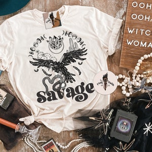 May include: A white t-shirt with a black and white graphic design of a raven, snake, and moon phases. The text "Savage" is printed in black on the shirt.