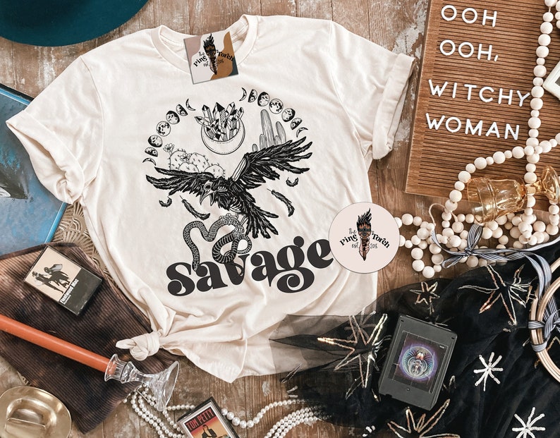 May include: A white t-shirt with a black and white graphic design of a raven, snake, and moon phases. The text "Savage" is printed in black on the shirt.
