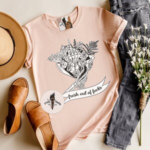 K&ouml;nnte beinhalten: Hellrosa T-Shirt mit einem schwarz-wei&szlig;en floralen Skelett-Hand-Design und dem Text "fresh out of fucks". Das Bild enth&auml;lt einen braunen Hut, Sandalen und Jeans.