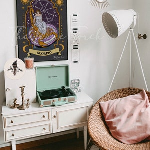 May include: A black and gold framed print of a zodiac sign, Scorpio, with a woman sitting on a scorpion. The print is hanging on a white wall with a gold eye mirror and a white lamp. A record player is on a white dresser with three drawers. A wicker chair with a pink pillow is in the corner of the room.