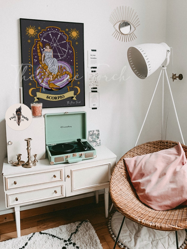 May include: A black and gold framed print of a zodiac sign, Scorpio, with a woman sitting on a scorpion. The print is hanging on a white wall with a gold eye mirror and a white lamp. A record player is on a white dresser with three drawers. A wicker chair with a pink pillow is in the corner of the room.