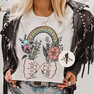 May include: White t-shirt with a rainbow, flowers, and two fists with the words "Girl Power" written on them. The text "The Pine Torch" is at the bottom of the shirt.
