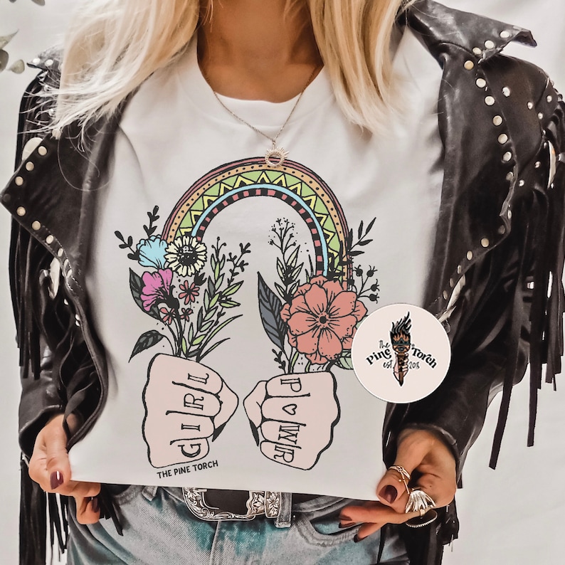 May include: White t-shirt with a rainbow, flowers, and two fists with the words "Girl Power" written on them. The text "The Pine Torch" is at the bottom of the shirt.