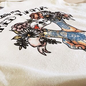 Pu&ograve; includere: Felpa color crema con una grafica raffigurante una figura incoronata di fiori con braccia tatuate. Il testo "I love my NAKED TAMA" &egrave; stampato sopra l'illustrazione. Il design incorpora fiori e un cuore.