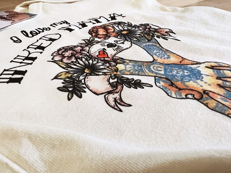 Pu&ograve; includere: Felpa color crema con una grafica raffigurante una figura incoronata di fiori con braccia tatuate. Il testo "I love my NAKED TAMA" &egrave; stampato sopra l'illustrazione. Il design incorpora fiori e un cuore.