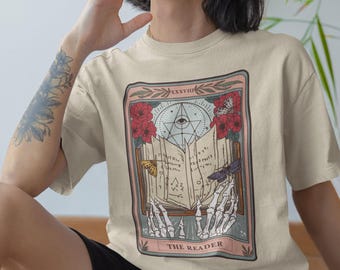 May include: A beige t-shirt with a tarot card design featuring a skeletal hand holding an open book with an all-seeing eye symbol. The card is titled "The Reader".