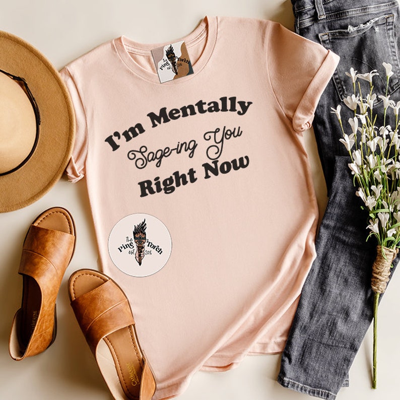 May include: A light pink t-shirt with the text "I'm Mentally Sage-ing You Right Now" in black. The shirt has a small logo on the bottom left corner that says "The Ring & Torch Co."