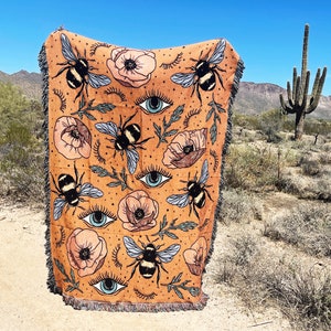 May include: A woven tapestry throw blanket with a mustard yellow background featuring a repeating pattern of bees, flowers, and stylized eyes.