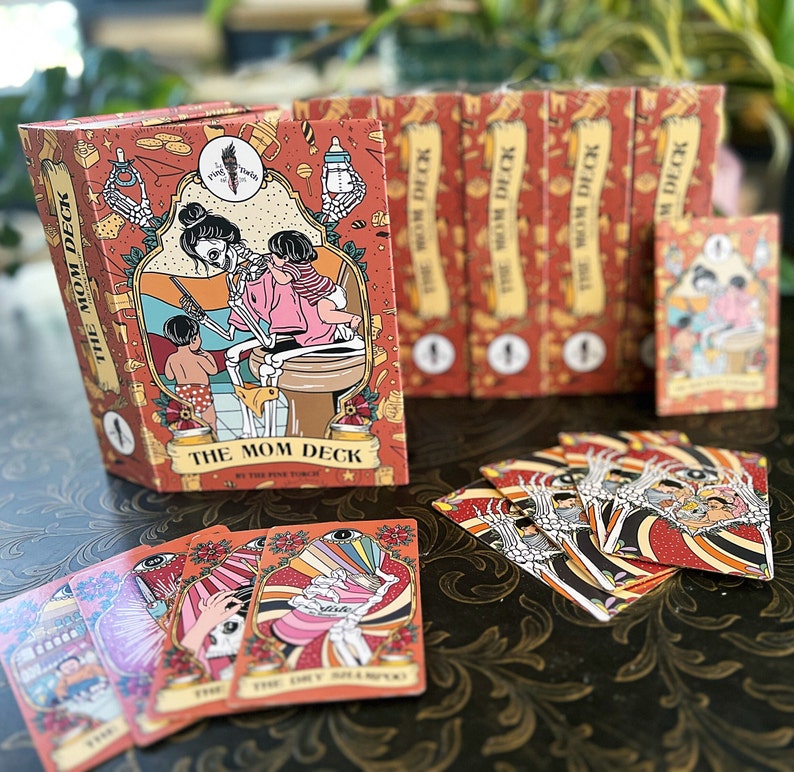 May include: A box of The Mom Deck playing cards with a colorful illustration of a skeleton mom and child on the front. The box is surrounded by a stack of other card decks.  The cards are laid out on a dark brown surface.