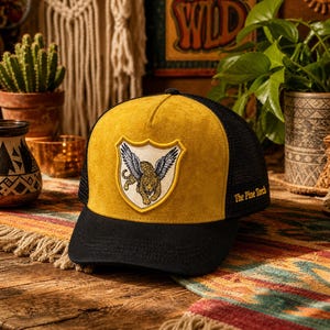 May include: A mustard yellow and black trucker hat featuring a shield-shaped patch on the front panel with an illustration of a winged leopard. The black mesh back panels include the embroidered text The Pine Torch in yellow thread.