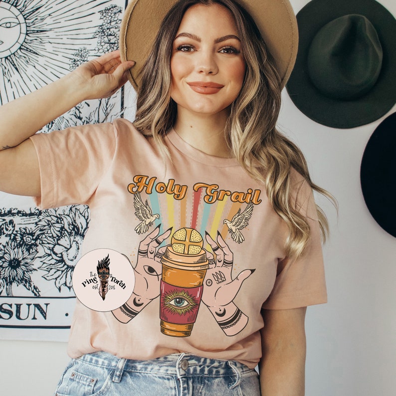 Op de afbeelding: Een perzikkleurig T-shirt met de tekst "Holy Grail" bedrukt op de voorkant. De afbeelding toont een koffiebeker met een oog en een kruis erop, omringd door handen en duiven.