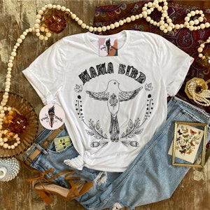 May include: White t-shirt with a black graphic of a bird with the text "Mama Bird" above it. The bird is surrounded by floral designs.