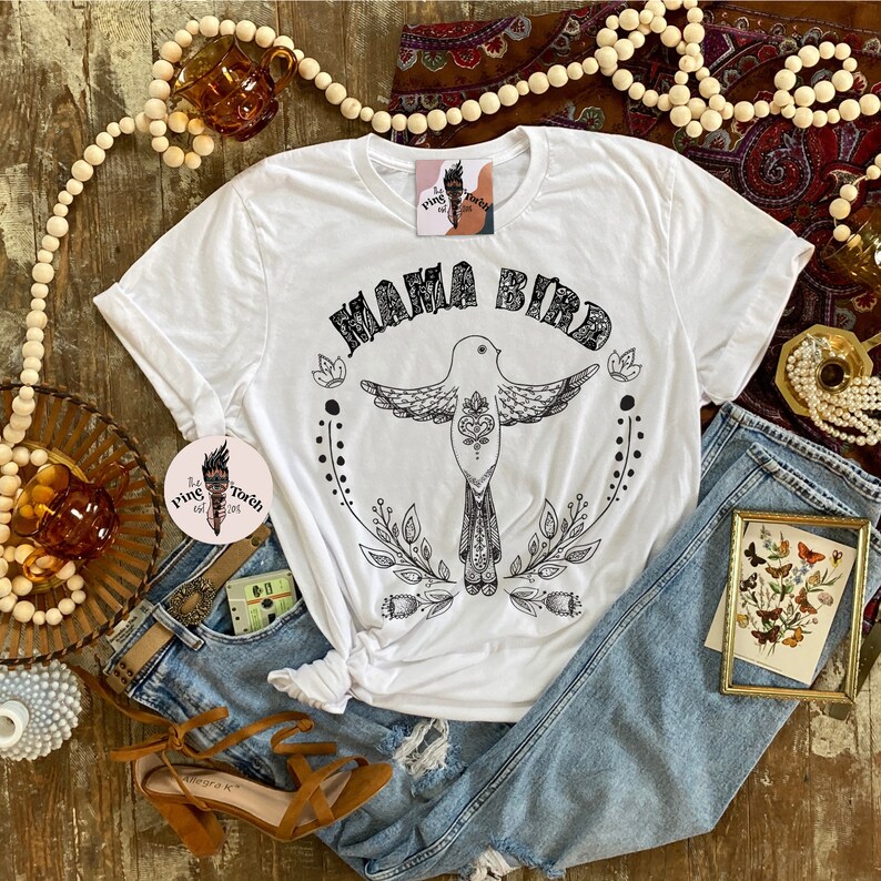 May include: White t-shirt with a black graphic of a bird with the text "Mama Bird" above it. The bird is surrounded by floral designs.