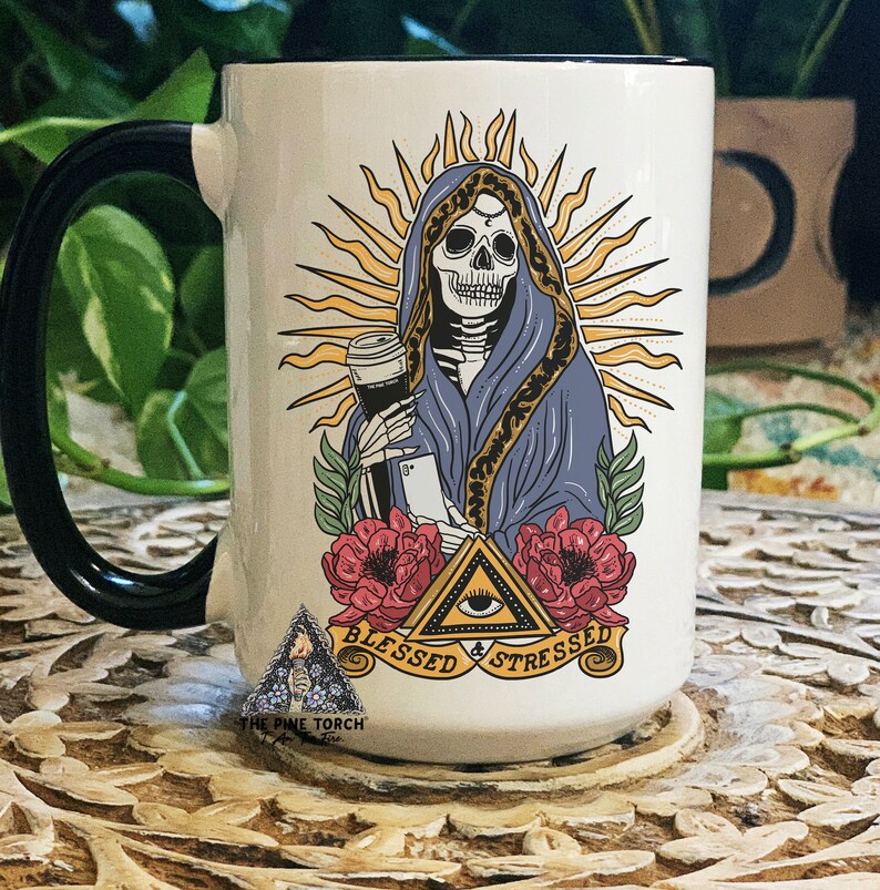 K&ouml;nnte beinhalten: Wei&szlig;er Keramikbecher mit schwarzem Henkel und Rand, mit einem Skelett in einem blauen Gewand, das eine Kaffeetasse und ein Telefon h&auml;lt. Das Design umfasst einen Sonnenaufgang, rote Blumen und den Text "Blessed & Stressed."