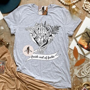 K&ouml;nnte beinhalten: Hellgraues T-Shirt mit einer schwarz-wei&szlig;en Grafik einer Skeletthand, die einen Blumenstrau&szlig; h&auml;lt. Das Banner darunter lautet "fresh out of fucks". Das Shirt hat kurze &Auml;rmel und einen Rundhalsausschnitt.