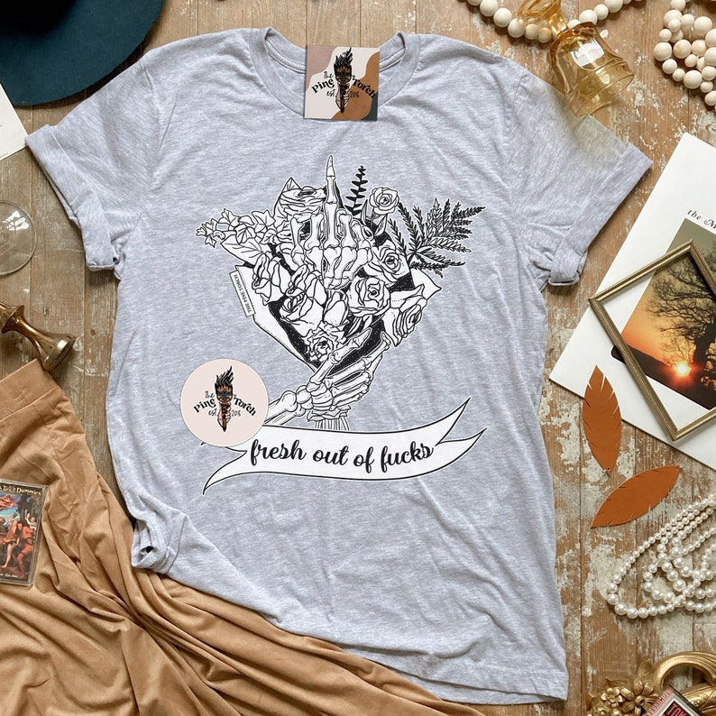 K&ouml;nnte beinhalten: Hellgraues T-Shirt mit einer schwarz-wei&szlig;en Grafik einer Skeletthand, die einen Blumenstrau&szlig; h&auml;lt. Das Banner darunter lautet "fresh out of fucks". Das Shirt hat kurze &Auml;rmel und einen Rundhalsausschnitt.