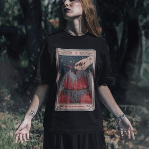 May include: Black t-shirt with a graphic print of a tarot card. The card is titled "The Burned Bridge" and features a hand reaching towards a burning bridge.