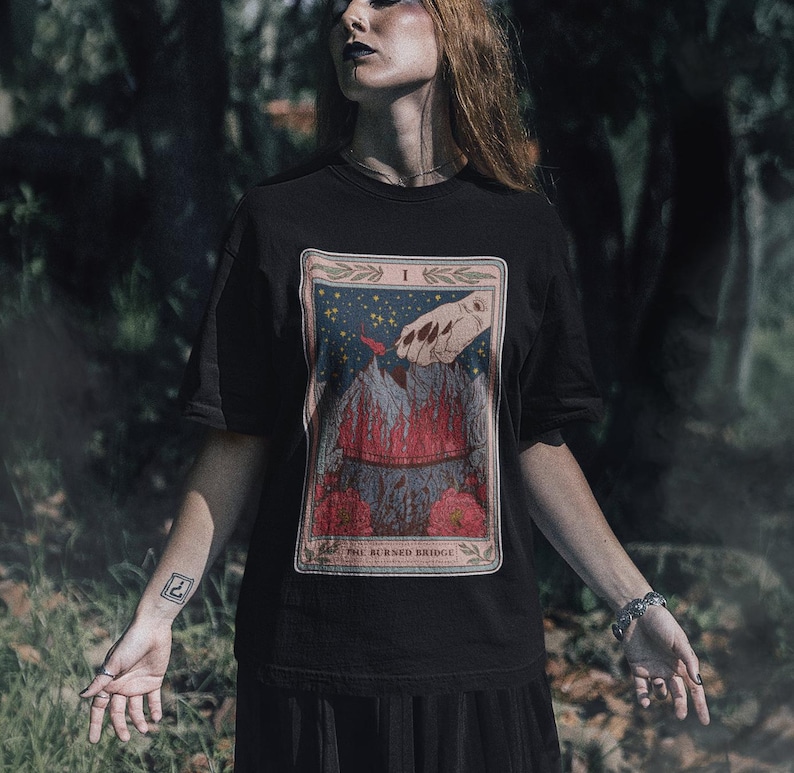 May include: Black t-shirt with a graphic print of a tarot card. The card is titled "The Burned Bridge" and features a hand reaching towards a burning bridge.