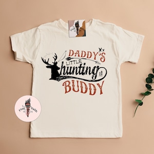 May include: Cream-colored t-shirt with the text "Daddy's Little Hunting Buddy" in red and black. The design includes a deer silhouette, bow and arrow, and tree graphics. A small logo is visible on the shirt.