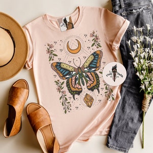 May include: A pink t-shirt with a graphic of a butterfly with eyes, a crescent moon, and a crystal. The butterfly is surrounded by flowers and stars. The text "The Pink Torch est. 2016" is on a sticker in the bottom right corner of the image.