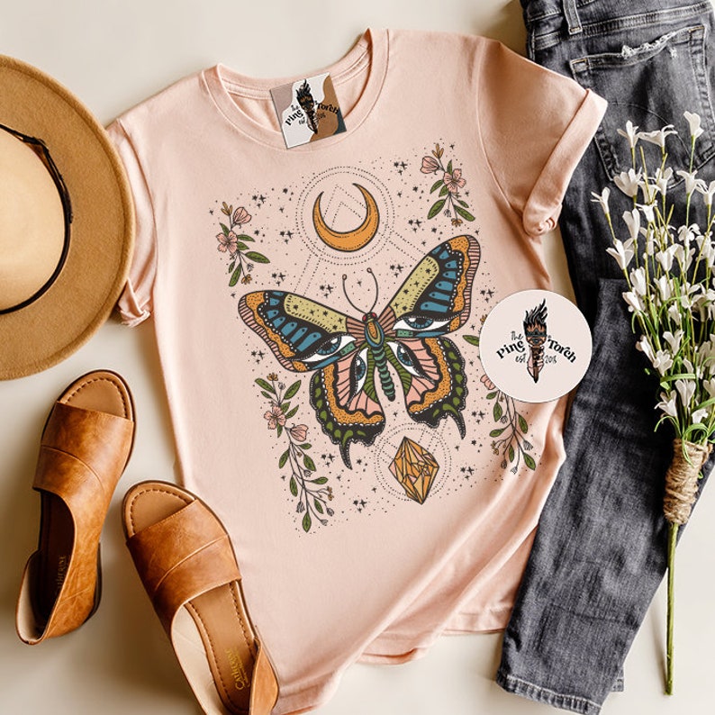 May include: A pink t-shirt with a graphic of a butterfly with eyes, a crescent moon, and a crystal. The butterfly is surrounded by flowers and stars. The text "The Pink Torch est. 2016" is on a sticker in the bottom right corner of the image.