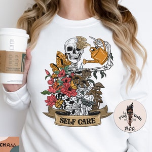 May include: White sweatshirt with a graphic of a skeleton watering flowers and mushrooms. The graphic has the text "Self Care" on a banner.