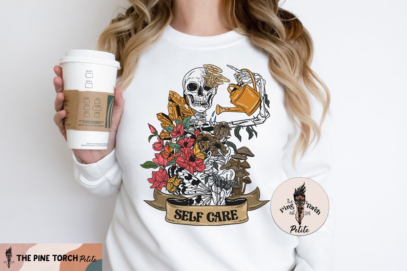 May include: White sweatshirt with a graphic of a skeleton watering flowers and mushrooms. The graphic has the text "Self Care" on a banner.