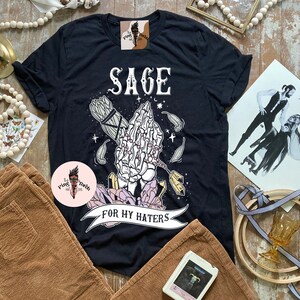 May include: Black t-shirt with a graphic of a skeleton hand holding a sage bundle and crystals. The text "Sage for my haters" is printed on the shirt.