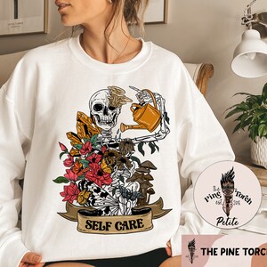 May include: White sweatshirt with a graphic of a skeleton watering a flower arrangement with crystals and mushrooms. The graphic has the text "SELF CARE" on a banner below it. The sweatshirt has a small logo on the bottom right corner that says "The Pine Torch Petite est. 2015".