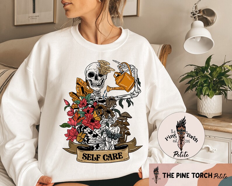 May include: White sweatshirt with a graphic of a skeleton watering a flower arrangement with crystals and mushrooms. The graphic has the text "SELF CARE" on a banner below it. The sweatshirt has a small logo on the bottom right corner that says "The Pine Torch Petite est. 2015".