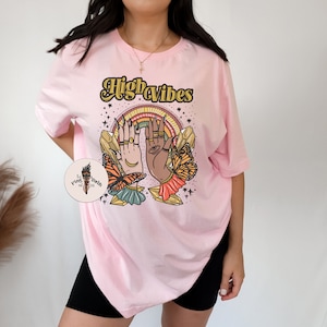 May include: Pink oversized t-shirt with the text "High Vibes" in a retro font. The graphic features two hands with crystals and butterflies.
