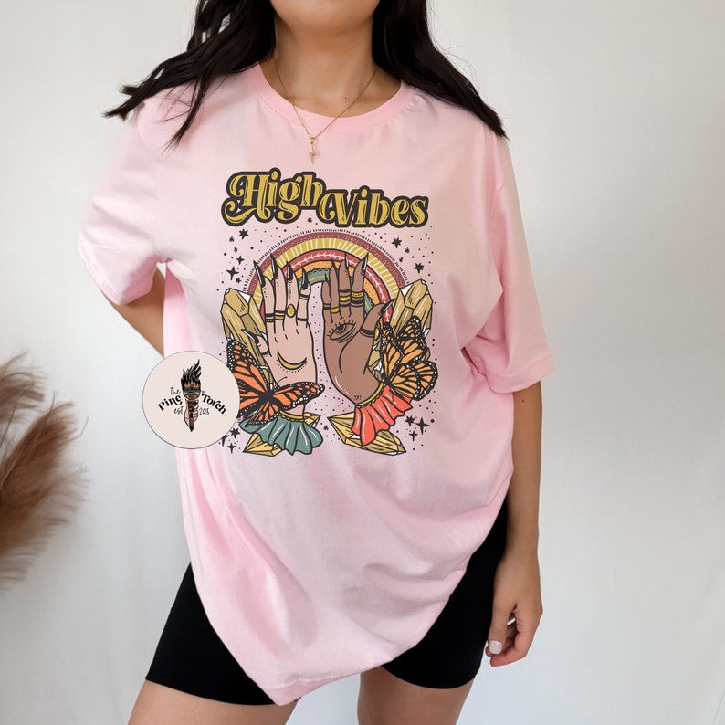 May include: Pink oversized t-shirt with the text "High Vibes" in a retro font. The graphic features two hands with crystals and butterflies.