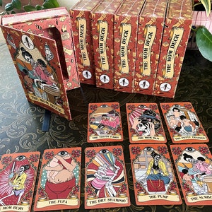 May include: A set of orange and red playing cards with illustrations of skeletons and the text "The Mom Deck". The cards are arranged in a fan shape on a brown patterned surface. The cards feature titles such as "The Tooth Fairy", "The Mom Bun", "The Potty Training", "The FUPA", "The Dry Shampoo", "The Pump", "The Number Two", and "The Mom Bush".