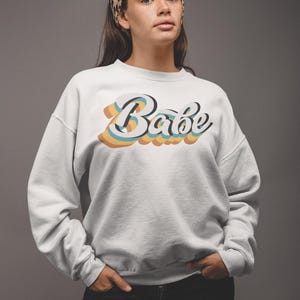 Może przedstawiać: Biała bluza z nadrukiem w stylu retro słowa "Babe" w kolorach tęczy.