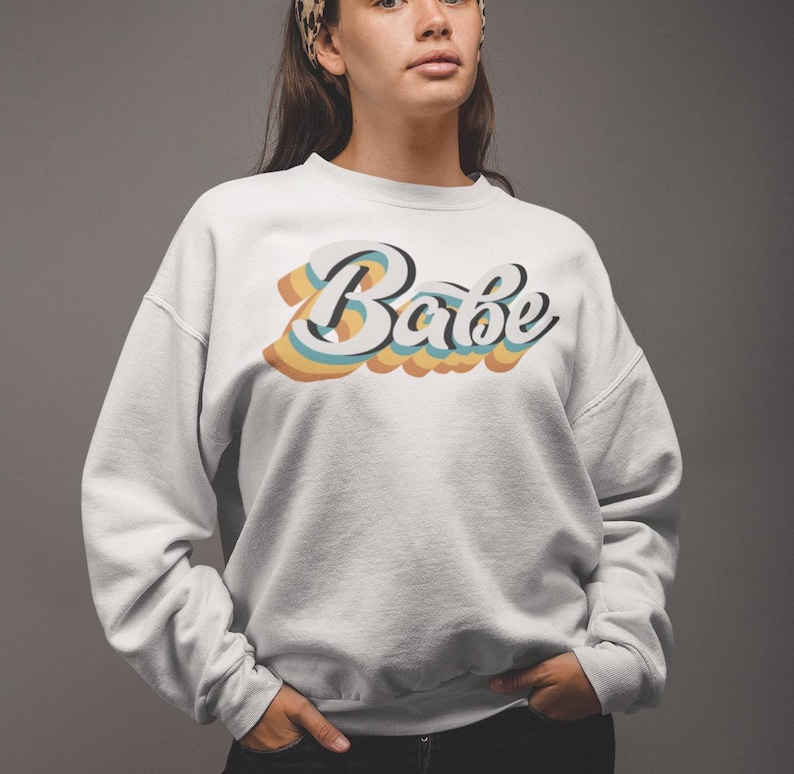 Może przedstawiać: Biała bluza z nadrukiem w stylu retro słowa "Babe" w kolorach tęczy.