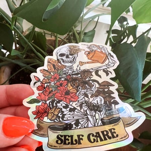 May include: A holographic sticker featuring a skeleton watering a plant with a watering can. The skeleton is surrounded by flowers, butterflies, and mushrooms. The sticker has a banner that reads "SELF CARE" in gold lettering.