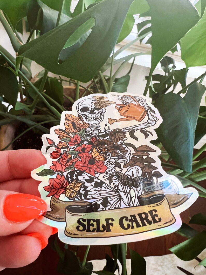 May include: A holographic sticker featuring a skeleton watering a plant with a watering can. The skeleton is surrounded by flowers, butterflies, and mushrooms. The sticker has a banner that reads "SELF CARE" in gold lettering.