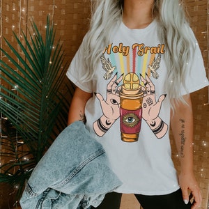 Op de afbeelding: Wit T-shirt met een afbeelding van twee handen die een koffiebeker vasthouden met een heilige graal erop. De tekst "Holy Grail" staat boven de afbeelding. De tekst "Je ne regrette rien" staat aan de rechterkant van het T-shirt.