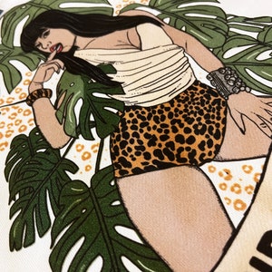 K&ouml;nnte beinhalten: Eine wei&szlig;e Tragetasche mit einer bunten Illustration einer Frau, die einen Leoparden-Badeanzug und ein wei&szlig;es Oberteil tr&auml;gt. Die Frau ist von gr&uuml;nen tropischen Bl&auml;ttern umgeben und der Text "Wild Noods" ist auf die Tasche gedruckt.