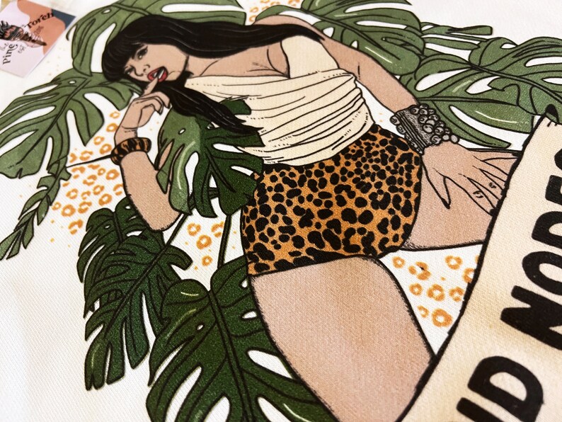 K&ouml;nnte beinhalten: Eine wei&szlig;e Tragetasche mit einer bunten Illustration einer Frau, die einen Leoparden-Badeanzug und ein wei&szlig;es Oberteil tr&auml;gt. Die Frau ist von gr&uuml;nen tropischen Bl&auml;ttern umgeben und der Text "Wild Noods" ist auf die Tasche gedruckt.