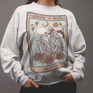 May include: White sweatshirt with a graphic print of a tarot card depicting two skeletons holding hands and flowers. The card is labeled "III THE LOVERS".