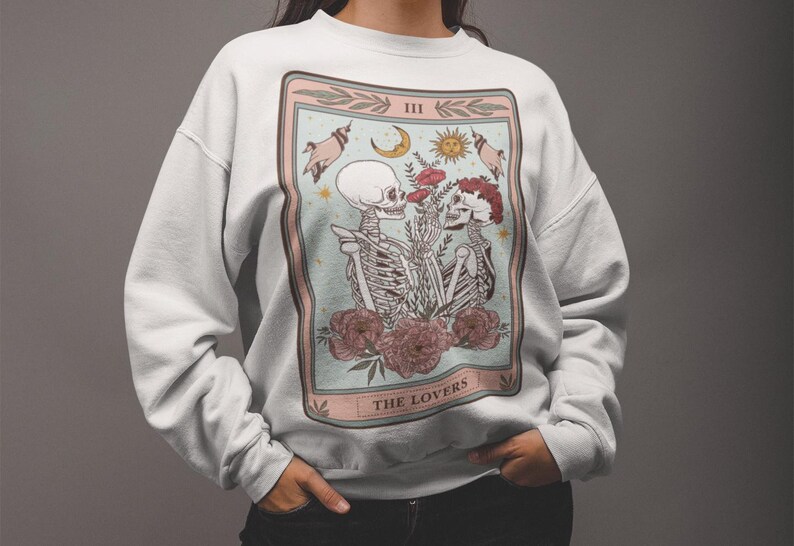 May include: White sweatshirt with a graphic print of a tarot card depicting two skeletons holding hands and flowers. The card is labeled "III THE LOVERS".