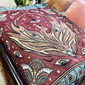 May include: A maroon tapestry throw blanket with a colorful, intricate design featuring a phoenix with multiple eyes and a paisley pattern. The blanket has a fringed edge.