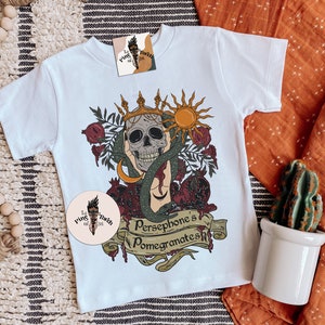 May include: White t-shirt featuring a graphic of a skull wearing a crown, surrounded by pomegranates and a snake. The text "Persephone's Pomegranates" is written on a banner below the skull.